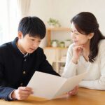学生服の中学生と母親が明るいダイニングで通知表を開いて話している様子