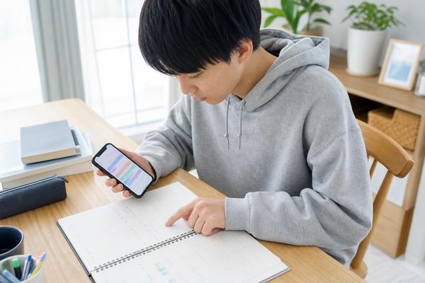 スマートフォンの勉強計画アプリと紙の予定表を見比べて1週間の勉強計画を立てている高校生