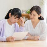 明るいダイニングテーブルで成績表を見て不安そうな中学生と、隣で穏やかに寄り添う母親