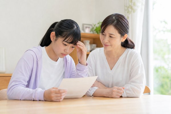 明るいダイニングテーブルで成績表を見て不安そうな中学生と、隣で穏やかに寄り添う母親