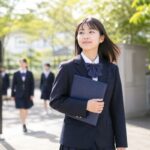 通知表のフォルダを抱えて明るい校門を歩く制服姿の中学生