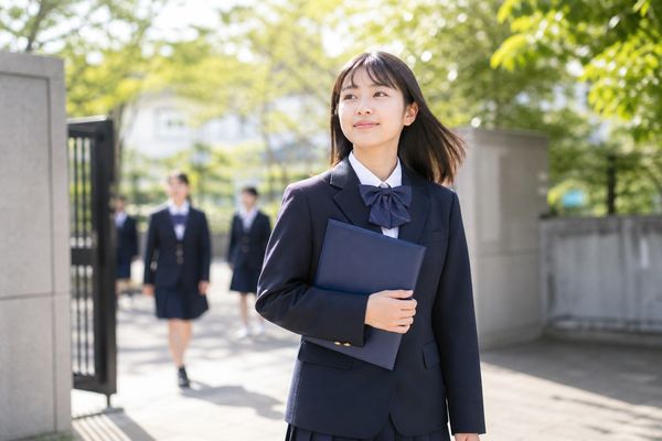 通知表のフォルダを抱えて明るい校門を歩く制服姿の中学生