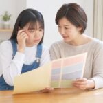 自宅で通知表を母親と一緒に見て考え込む中学生の女の子