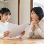 通知表を見て笑顔になる中学生と、隣で一緒に喜ぶ母親の様子