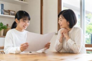 通知表を見て笑顔になる中学生と、隣で一緒に喜ぶ母親の様子