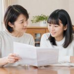 母親と中学生の娘が、明るいダイニングで通知表を広げて落ち着いて話し合っている様子