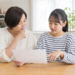明るいダイニングで母親と中学生の娘が並んで座り、成績表を一緒に見ながら穏やかに話している様子
