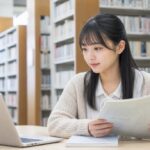 図書館でノートパソコンと問題冊子を見比べて確認する女子高校生
