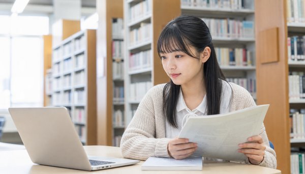 図書館でノートパソコンと問題冊子を見比べて確認する女子高校生
