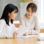 明るいダイニングテーブルで、私服の母親と中学1年生の娘が並んで座り、返却された答案用紙とノートを一緒に見ながら穏やかに話している様子