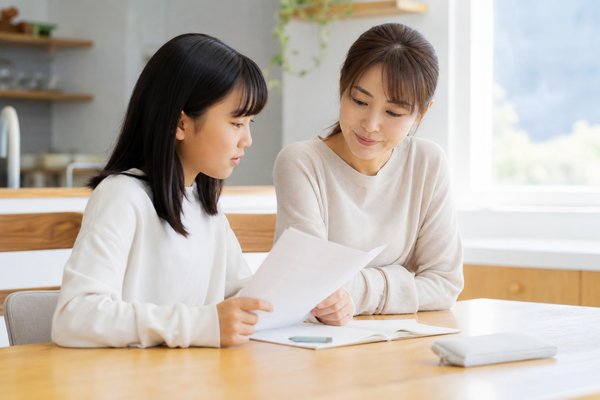 明るいダイニングテーブルで、私服の母親と中学1年生の娘が並んで座り、返却された答案用紙とノートを一緒に見ながら穏やかに話している様子