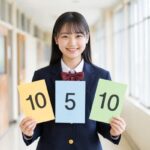 明るい高校の廊下で、制服姿の女子高校生が「10」「5」「10」と書かれた3枚の色分けカードを並べて持ち、正面に向かって笑顔で立っている様子
