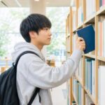 明るい図書館の書棚で、私服の高校生が自分のレベルに合う数学の問題集を1冊選び出している様子