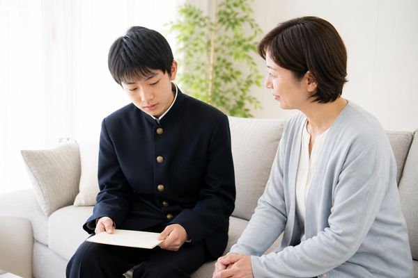 学生服姿の中学生が通知表を見つめ、隣に座る保護者が静かに話しかけている様子