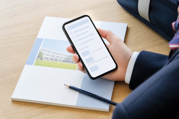 制服姿の高校生が机の学校案内の上でスマートフォンを持ち、模試の正式名称を検索して確かめている手元