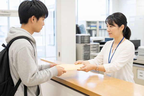 学校の事務窓口で高校生が封緘された成績証明書の封筒を職員から受け取る場面