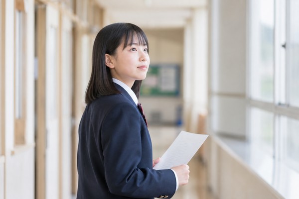 日中の明るい中学校の廊下で、紺のブレザー制服を着た女子生徒がテスト用紙を手に持ち、窓の方へ視線を向けて考えている様子