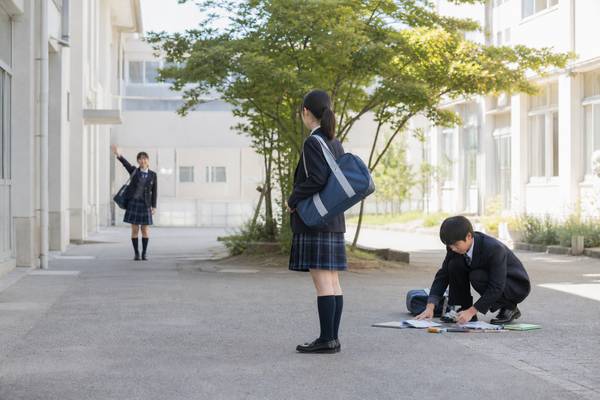 校庭で、手を振る友達と落とし物を拾う同級生の間に立ち止まる制服の女子中学生