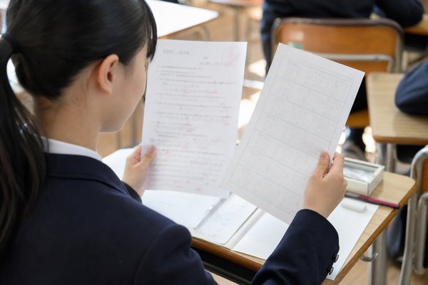 教室の席で、返却された定期テストの答案と観点別の記録用紙を見比べている制服姿の中学生