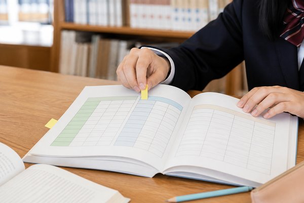 図書室の木の机で、制服の生徒が学校資料の見開きに載った換算表の一行に黄色い付箋を貼って印をつけている手元の様子