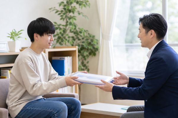 中学生が明るい教室で、たまっていた提出物のファイルを先生へ手渡している様子