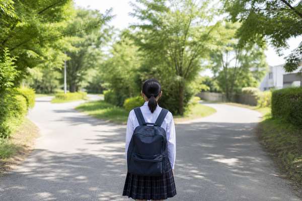 制服姿の中学生が通学路の分かれ道に立ち、どちらの道も明るく開けているのを眺めている後ろ姿