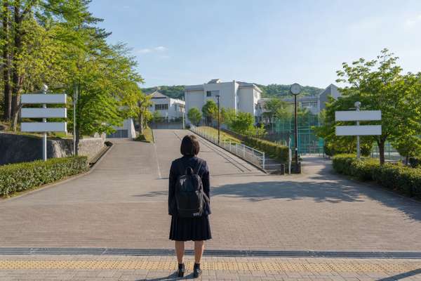 リュックを背負った中学生が分かれ道に立ち、先に見える高校の校舎を眺めている後ろ姿
