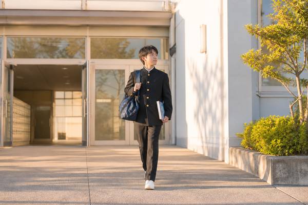 学ラン姿の中学3年生の男子が、部活動のバッグと教科書を持って夕方の校舎から歩き出す様子