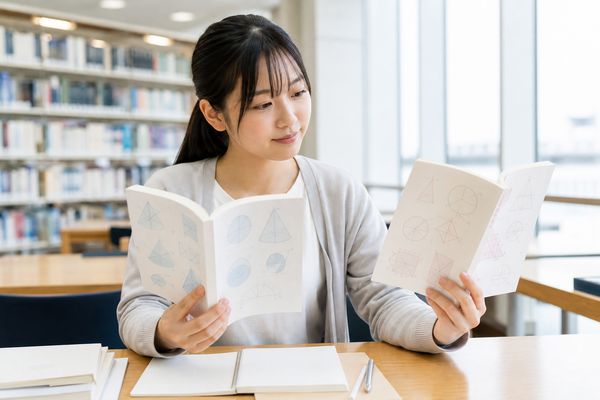 大学図書館風の自習スペースで、私服の高校生が分野別教材と総合問題集の2冊を見比べて選んでいる場面