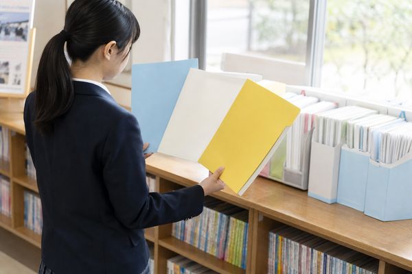 高校の進路資料コーナーで制服の生徒が3種類の出願資料を手に取って見比べている様子