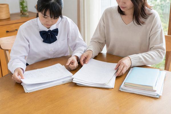 制服姿の中学生と保護者が、返ってきた答案やワークをダイニングテーブルに広げ、三つの束に仕分けている様子