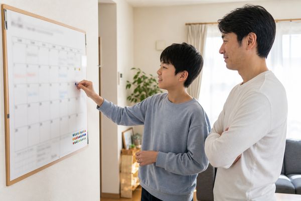 父親と中学生の息子がリビングの壁掛けカレンダーの前に立ち、次にやることへシールを貼りながら相談している様子