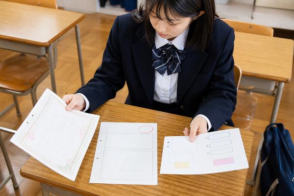 放課後の教室の机で、制服の女子高校生が手書きのレポート、図形の書かれたテスト答案、発表用のカードの3種類の資料を並べて見比べている様子
