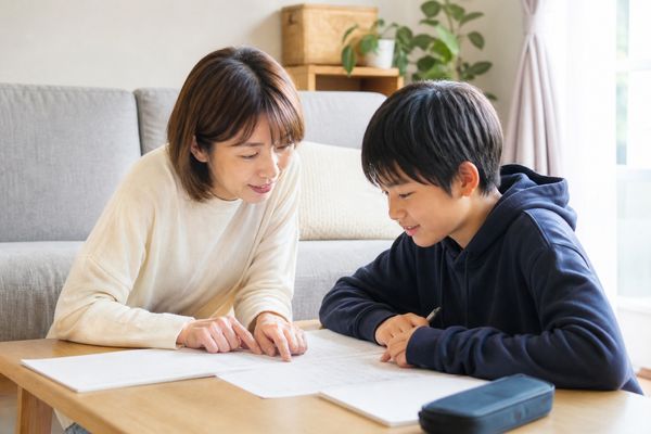 明るいリビングのローテーブルで、私服の母親と中学1年生の男子が学校のプリントをのぞき込み、母親が紙面を指さしながら一緒に確認している様子