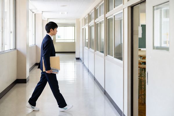 制服姿の中学生が筆記用具と書類を持って、明るい校舎の廊下を試験教室へ向かって歩いている様子