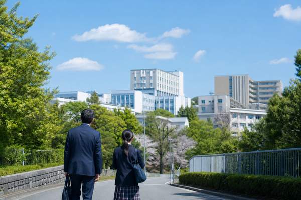青空の下で制服の中学生と父親が複数の高校校舎へ続く道を歩く後ろ姿