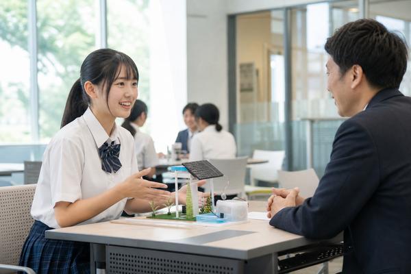制服姿の高校生が大学の面談室で自分の研究作品を見せながら大学の先生に説明している様子