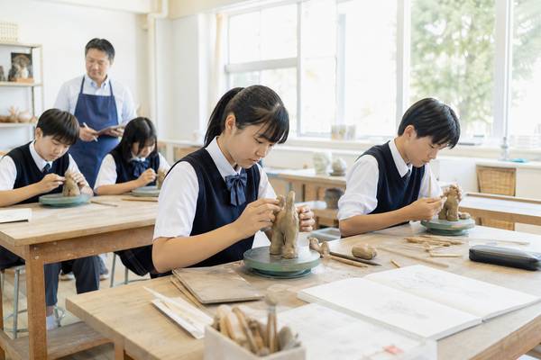 中学校の美術室で制服姿の生徒たちが粘土の作品を制作し、教員が机の間を回っている様子