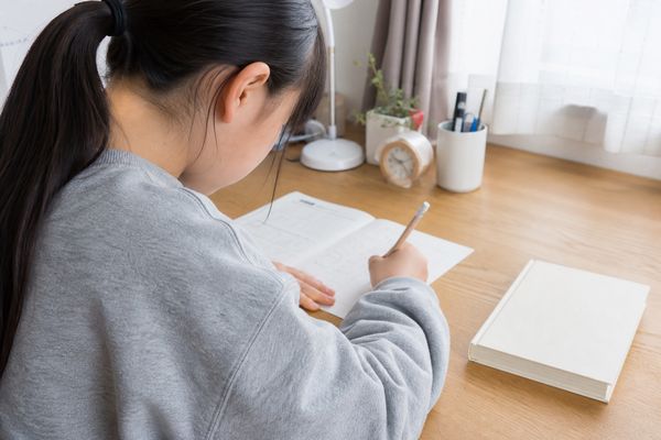 明るい自室の机で、私服の中学1年生の女子生徒が参考書を閉じたまま、鉛筆で問題プリントを自力で解いている様子