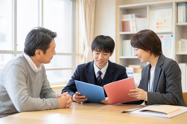 明るい進路相談室で、制服姿の男子高校生と保護者が先生と2冊の資料を分けて確認している様子