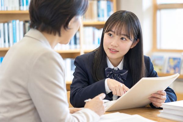 明るい学校図書室で、制服姿の女子高生が資料の一箇所を指しながら先生に質問している様子