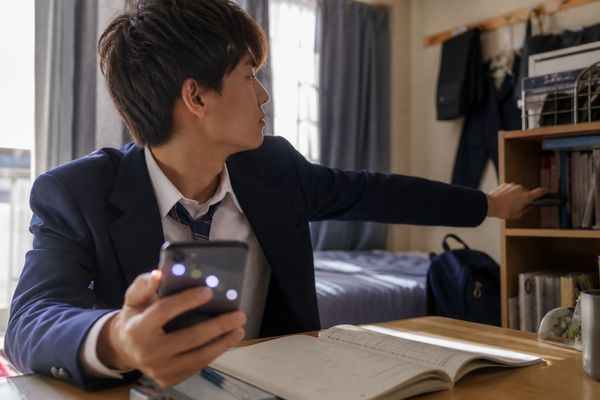 通知が並ぶスマートフォンを手にした制服の高校生が机から離れた棚へスマホを置こうとしている様子
