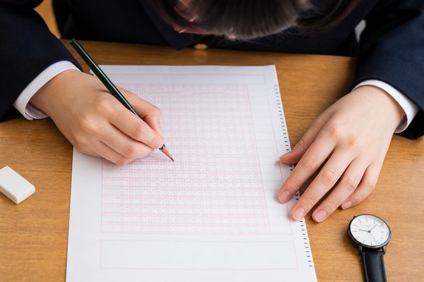 試験机の上で、鉛筆を持った手が解答欄を一行ずつ指でたどって見直している手元の接写