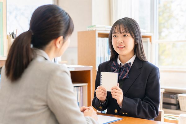 質問メモを手に進路指導室で先生へ相談する高校生の女子生徒