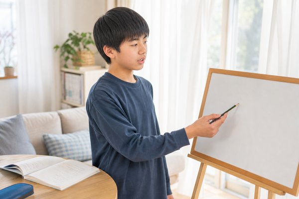 自宅リビングで、私服の男子中学生が小さなホワイトボードの前に立ち、一問の解き方を声に出して自分で説明している様子