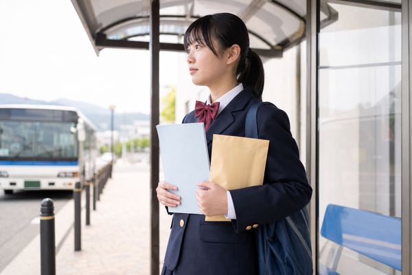 制服姿の中学生が明るいバス停で成績のファイルと模試の封筒を抱えて考えている様子
