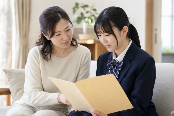 明るいリビングのソファで、母親と制服姿の高校生の娘が並んで座り、成績資料のファイルを一緒に開いて穏やかに話している場面