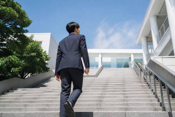 青空の下、制服姿の高校生がノートを抱えて校舎へ続く階段を自分の足で上っていく後ろ姿
