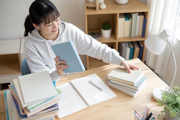 明るい自宅の机で、私服の高校生が積み上がった参考書から使う一冊を選び、残りを脇へ寄せている様子