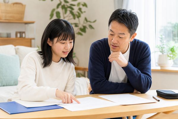 明るいリビングのテーブルで、私服の父親と中学1年生の娘が並んで座り、学校のプリントを前に娘が項目を指さしながら二人で計画を立てている様子